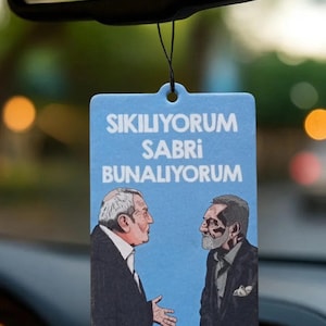 May include: A light blue car air freshener with the text "SIKILIYORUM SABRI BUNALIYORUM" and illustrations of two men in suits. The air freshener is hanging from a black string.