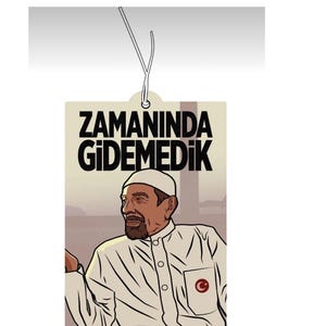 May include: A beige car air freshener with a black and white illustration of a man wearing a white hat and a white shirt with a red circle and a white crescent moon on the pocket. The text "ZAMANINDA GİDEMEDİK" is printed in black at the top of the air freshener.
