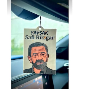 May include: A car air freshener with a cartoon illustration of a man with a beard and the text "YAVSAK Safi Rüzgar" on a white background.