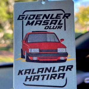 May include: A light blue car air freshener with a red car graphic. The air freshener has the text "Gidenler Masal Olur" and "Kalanlar Hatira" in black, stylized font. A white string is attached at the top for hanging.
