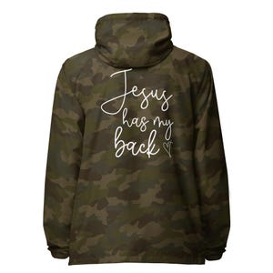 May include: A camouflage jacket with a hood. The back of the jacket displays the white cursive text "Jesus has my back" with a small heart and cross symbol. The jacket is in shades of green, brown, and black.