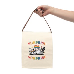 May include: A cream-colored canvas bag with a brown strap featuring a graphic of a raccoon wearing rainbow heart-shaped sunglasses, holding a rainbow flag, and surrounded by colorful stars. The text SURPRISE is printed above and below the raccoon.