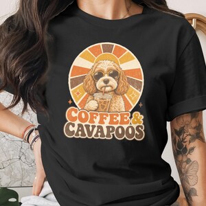 May include: A black short-sleeved t-shirt featuring a graphic of a curly-haired dog wearing sunglasses and drinking iced coffee. Behind the dog is a retro-style sunburst design in orange and yellow. The text COFFEE & CAVAPOOS is printed below.