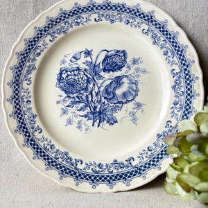 Può includere: Un piatto in ceramica bianca con un motivo floreale blu. Il piatto presenta un bordo smerlato e un bordo dettagliato con un motivo ripetuto. Il centro del piatto mostra un bouquet floreale blu.