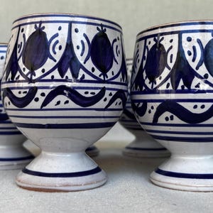 Può includere: Un set di cinque calici in ceramica con base bianca e motivi blu scuro dipinti a mano. Ogni calice presenta una coppa arrotondata, un gambo corto a piedistallo e una base circolare. Il design include motivi floreali e linee ondulate.