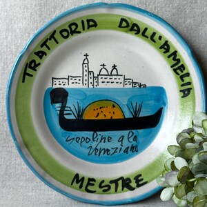 May include: A hand-painted ceramic plate with a blue, green, and white design. The plate features a gondola, a cityscape, and the text "Trattoria Dall'Amelia Mestre" in black lettering. The plate has a scalloped edge.