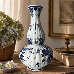 Könnte beinhalten: Eine hohe, weiße Keramikvase mit geriffelter Doppel-Kürbis-Form. Die Oberfläche ist mit komplexen blauen Blumenmustern und feinen Linien verziert. Die Vase hat einen schmalen Hals, der sich zu einem runden Boden hin verbreitert, und zeigt eine klassische blau-weiße Farbpalette mit glänzender Oberfläche.