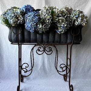 May include: A rectangular, dark grey, fluted metal planter box filled with a dense arrangement of blue and pale green hydrangea flowers, supported by a tall, brown, wrought iron stand featuring decorative scrollwork and four curved legs.
