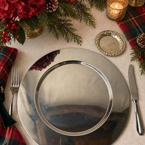 May include: A large, circular, polished silver-tone metal charger plate with a smooth, reflective surface. The plate features a wide, flat rim and a slightly recessed center, providing a clean, metallic aesthetic for a formal table setting.