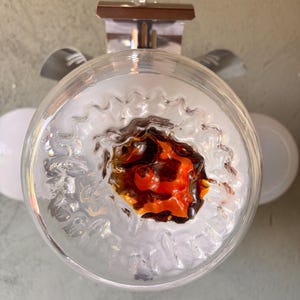 Puede incluir: Una lámpara circular de cristal con un borde exterior transparente y ondulado, y un núcleo texturizado con tonos arremolinados de ámbar, naranja y marrón oscuro. La lámpara está suspendida por un soporte de metal plateado con cadena.