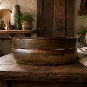 Puede incluir: Una gran palangana de cobre vintage, redonda, con una pátina oscura y desgastada y oxidación visible. El recipiente presenta un cuerpo ancho ligeramente curvado, una base plana y dos pequeñas asas de metal unidas a los lados con textura.