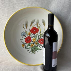 Può includere: Una grande ciotola rotonda in ceramica color bianco sporco con un bordo giallo brillante. L'interno presenta una colorata illustrazione botanica di papaveri rossi, fiori di campo gialli e spighe di grano. Una bottiglia di vino scura è posta davanti.
