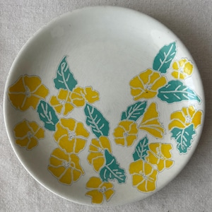 Può includere: Un piatto rotondo in ceramica con base bianca caratterizzato da un motivo ripetuto di fiori giallo brillante e foglie verde acqua. Il design floreale è distribuito sulla superficie, mostrando contorni decisi e una finitura liscia e lucida.