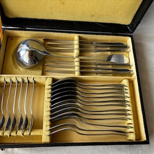 May include: A silver-toned stainless steel flatware set housed in a black rectangular storage case with a yellow interior lining. The set includes a large soup ladle, several dinner knives, and a collection of matching forks and spoons.