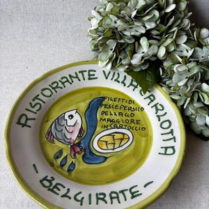 Può includere: Piatto in ceramica dipinto a mano con un disegno di pesce e testo in italiano. Il bordo è verde e il centro è giallo. Il piatto reca la scritta "Ristorante Villa Carlotta - Belgirate".