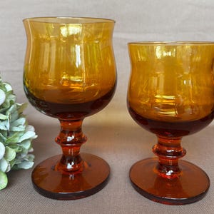 Puede incluir: Dos copas de cristal de color ámbar con forma de cuenco liso y redondeado y un tallo grueso de varios niveles que conduce a una base circular. El cristal presenta un degradado de naranja intenso en la base a un amarillo dorado claro.