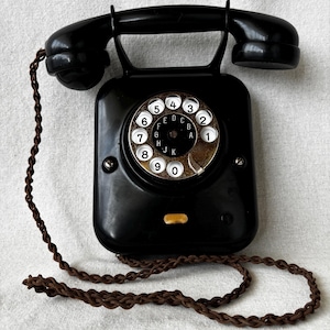 Può includere: Un telefono a disco nero con una cornetta appoggiata sopra. Il quadrante circolare presenta numeri bianchi da uno a zero e lettere dalla A alla K al centro. Un cavo in tessuto intrecciato marrone è fissato al lato della base nera lucida.