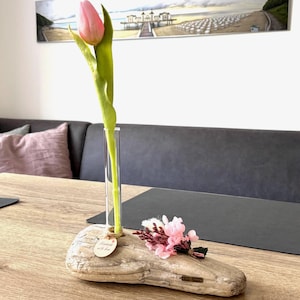 Könnte beinhalten: Eine rosa Tulpe in einer klaren Glasvase, auf einem Stück Treibholz. Der Treibholzsockel hat ein kleines Etikett mit der Aufschrift "Handmade with love". Ein kleines Bündel rosa Blumen ist befestigt. Die Vase steht auf einem Holztisch.