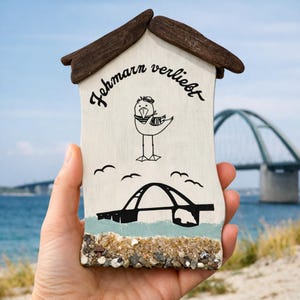 Puede incluir: Placa decorativa de madera con forma de casa, con tejado marrón y cuerpo blanco. La placa presenta una gaviota de dibujos animados, una ilustración de puente y el texto "Fehmarn verliebt" en negro. La base está decorada con piedrecitas y arena.