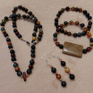 May include: A jewelry set featuring a necklace, bracelet, and earrings. The necklace and bracelet are made of dark, round beads in shades of black, brown, and green, with gold-colored spacers. The earrings have a similar design, with a rectangular pendant on the bracelet.
