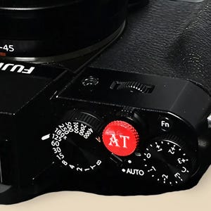 Könnte beinhalten: Nahaufnahme eines schwarzen Kameragehäuses mit verschiedenen Drehknöpfen und Tasten. Ein roter Drehknopf mit der Aufschrift "AT" ist prominent, zusammen mit einem Drehknopf für die Verschlusszeiteinstellungen. Die Kamera hat einen strukturierten Griff.