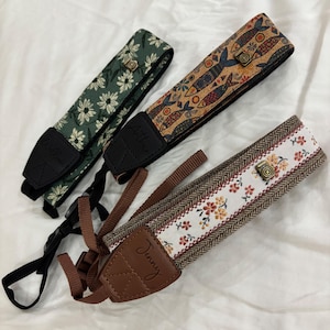 Könnte beinhalten: Drei Kameragurte mit verschiedenen Mustern. Einer ist grün mit weißen Blumen, ein anderer hat ein Fischmuster auf Korkbasis, und der dritte hat ein Blumen- und Fischgrätenmuster. Jeder Gurt hat ein Lederende mit einem Namen.