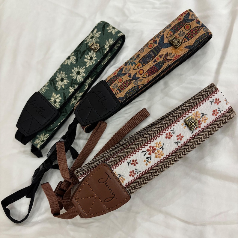 May include: Three camera straps with unique designs. One strap features a green floral pattern, another has a cork-like design with fish, and the third has a floral and herringbone pattern. Each strap has a leather end piece with a name.