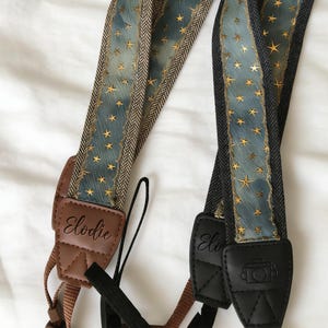 May include: Two camera straps with a celestial design. The straps feature a blue background with gold stars and a brown and black herringbone pattern. The leather ends are brown and black, with the name "Elodie" embossed on one strap.