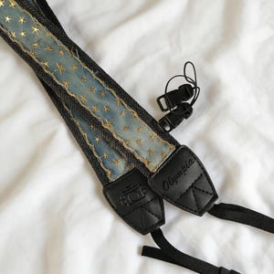 May include: A denim camera strap with gold star embellishments. The strap has black leather ends with the word "Olympia" and a camera icon. Includes black plastic clips for attachment.