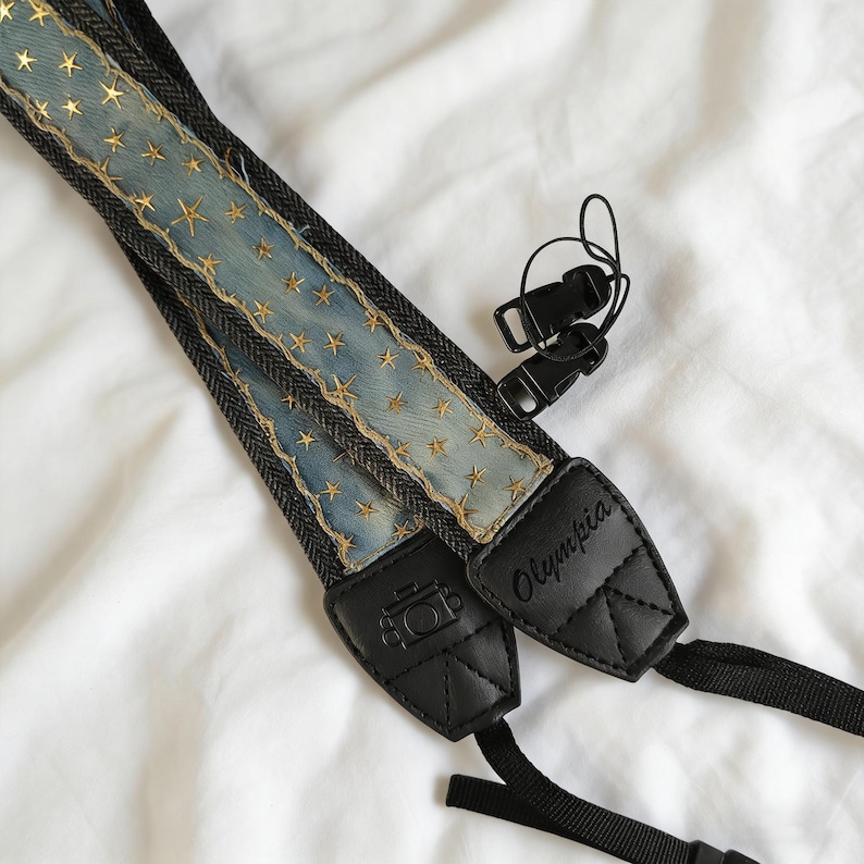 May include: A denim camera strap with gold star embellishments. The strap has black leather ends with the word "Olympia" and a camera icon. Includes black plastic clips for attachment.