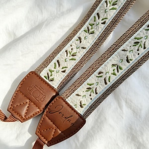 Könnte beinhalten: Zwei Kameragurte mit braunen Lederenden und einer gewebten Mitte. Die Mitte hat einen weißen Hintergrund mit grüner und brauner Blumenstickerei. Die Lederenden sind mit einem Kamera-Symbol und dem Namen "Jordan" geprägt.