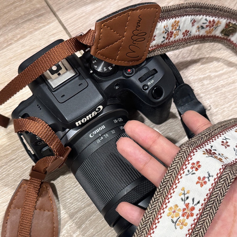 May include: A black Canon camera with a brown leather and floral patterned camera strap. The strap has the name "mailliiW" printed on the leather. The camera lens is black with the Canon logo.