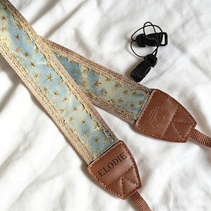 May include: A camera strap with a light blue denim-like pattern, adorned with gold star embroidery. The strap has brown leather ends with the name "ELODIE" and a logo. A black plastic clip is also included.