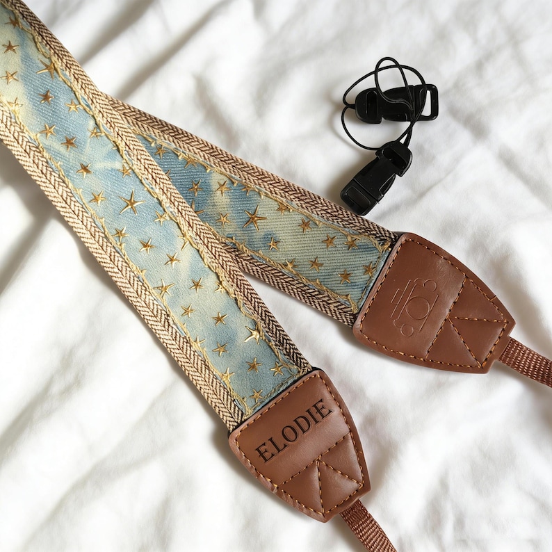 May include: A camera strap with a light blue denim-like pattern, adorned with gold star embroidery. The strap has brown leather ends with the name "ELODIE" and a logo. A black plastic clip is also included.