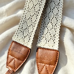 May include: A camera strap featuring a cream-coloured lace fabric with a scalloped edge. The strap ends are made of brown leather with triangular stitching. One leather end is embossed with the name Theodore, and the other features a camera icon.