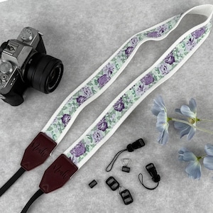 May include: A camera strap with a floral design of purple roses and green leaves on a white band. The strap has brown leather ends with the name "Noah" embossed. Includes black strap connectors and a camera.