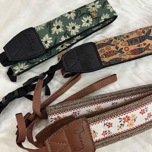 May include: Three camera straps with different patterns. One is green with white daisy-like flowers, another has a cork-like design with fish, and the third has a floral and herringbone pattern. Each strap has a black leather piece with a name.