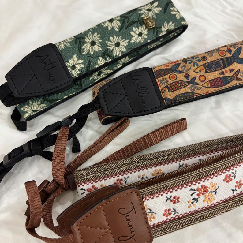 May include: Three camera straps with different patterns. One is green with white daisy-like flowers, another has a cork-like design with fish, and the third has a floral and herringbone pattern. Each strap has a black leather piece with a name.