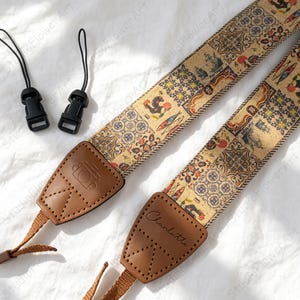 May include: A camera strap featuring a beige cork-like material with a multi-coloured tile and sardine pattern. It includes brown leather end tabs, one embossed with a camera icon and the other with the name Charlotte, plus two black plastic clips.