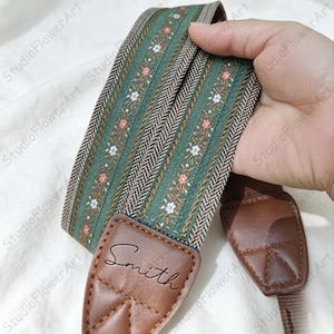 May include: A green and brown patterned guitar strap with floral accents and a brown leather end piece. The strap has the name "Smith" embossed on the leather. The strap is designed for a guitar.