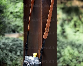 May include: A brown leather camera strap with the name "Genny" printed on it. The strap is attached to a silver and black camera with a black lens. A small yellow duck is attached to the camera. The background is blurred green foliage.