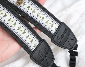 May include: A black camera strap featuring a white fabric center panel with a green floral pattern and small dark blue dots. The strap includes black leather-like end tabs, one embossed with a camera icon and the other with the name Emma.