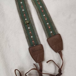 May include: Green and brown camera strap with a floral pattern and leather accents. The strap features a small camera-shaped button and the name "Alexander" on one of the leather pieces. The strap is designed to be worn around the neck.