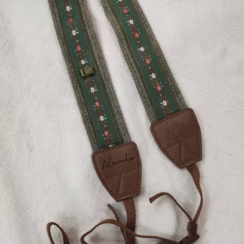 May include: Green and brown camera strap with a floral pattern and leather accents. The strap features a small camera-shaped button and the name "Alexander" on one of the leather pieces. The strap is designed to be worn around the neck.