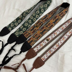 May include: Four patterned camera straps. One strap is dark green with white daisy flowers. Another is brown with fish designs. The third is white with floral patterns and brown trim. The fourth is green with white daisy flowers.
