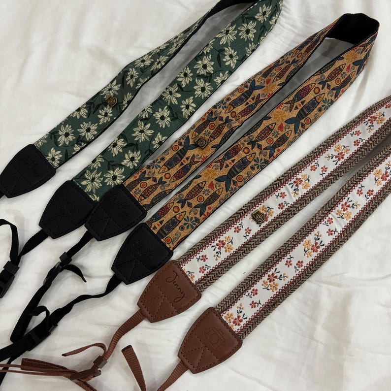 May include: Four patterned camera straps. One strap is dark green with white daisy flowers. Another is brown with fish designs. The third is white with floral patterns and brown trim. The fourth is green with white daisy flowers.