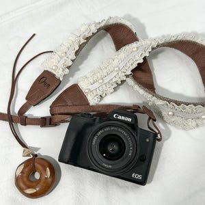 May include: A black Canon EOS camera with a lens, alongside a brown and white lace camera strap decorated with pearls. A brown leather strap and a donut-shaped pendant are also visible.