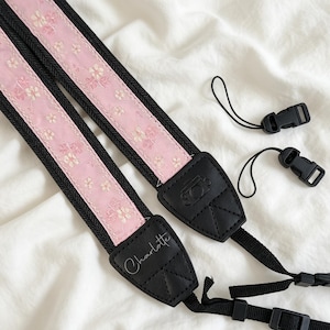May include: A camera strap featuring a light pink fabric centre with a repeating white and pink floral pattern, bordered by black webbing. The strap includes black leather-like end tabs, one embossed with a camera icon and one personalised with the name Charlotte.