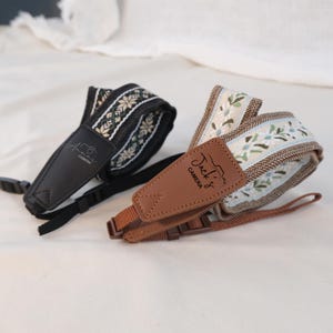 May include: Two camera straps with leather-like end tabs. One strap is black with a dark floral pattern and the text "Anna's CAMERA". The other is light beige with a blue and green floral pattern and the text "Jack's CAMERA" on a brown tab.