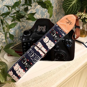 May include: A navy blue camera strap with a repeating pattern of white and black cat illustrations and red accents. The strap has a tan leather end piece with the word "Artisan" printed on it. The strap is attached to a black DSLR camera.